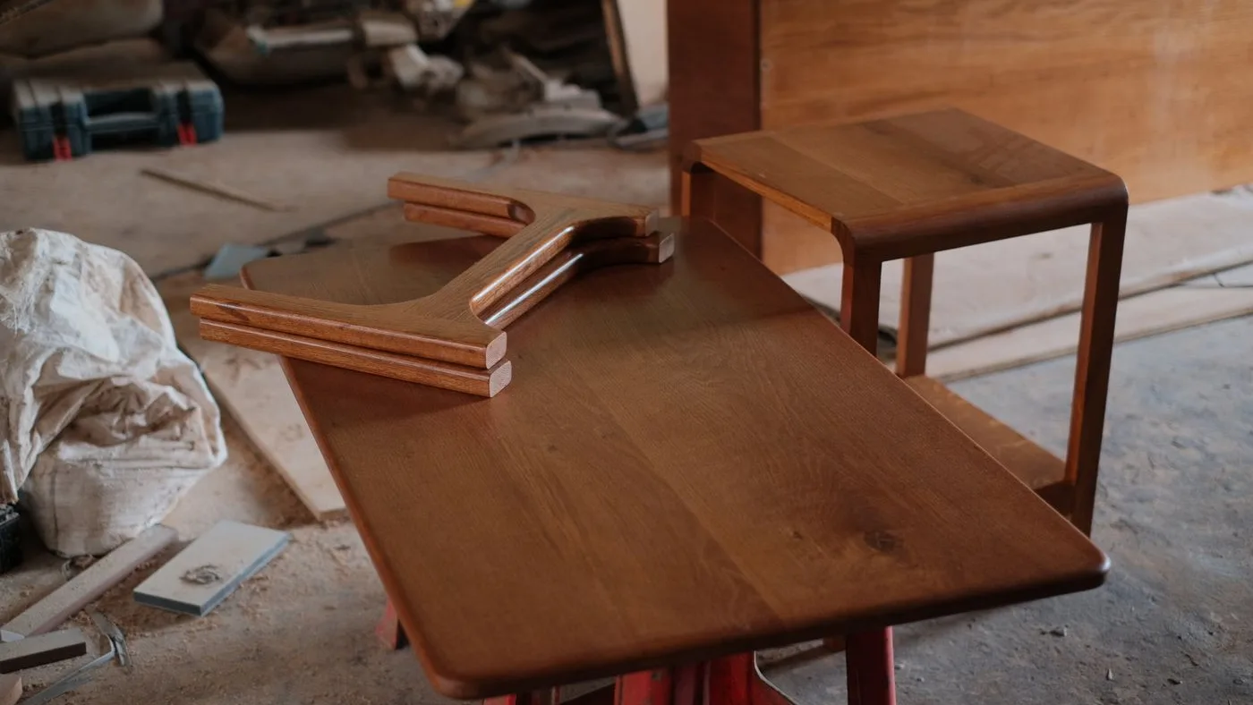 Stack of curved timber panels resting on a polished wooden table top beside a part-built side table in a workshop
