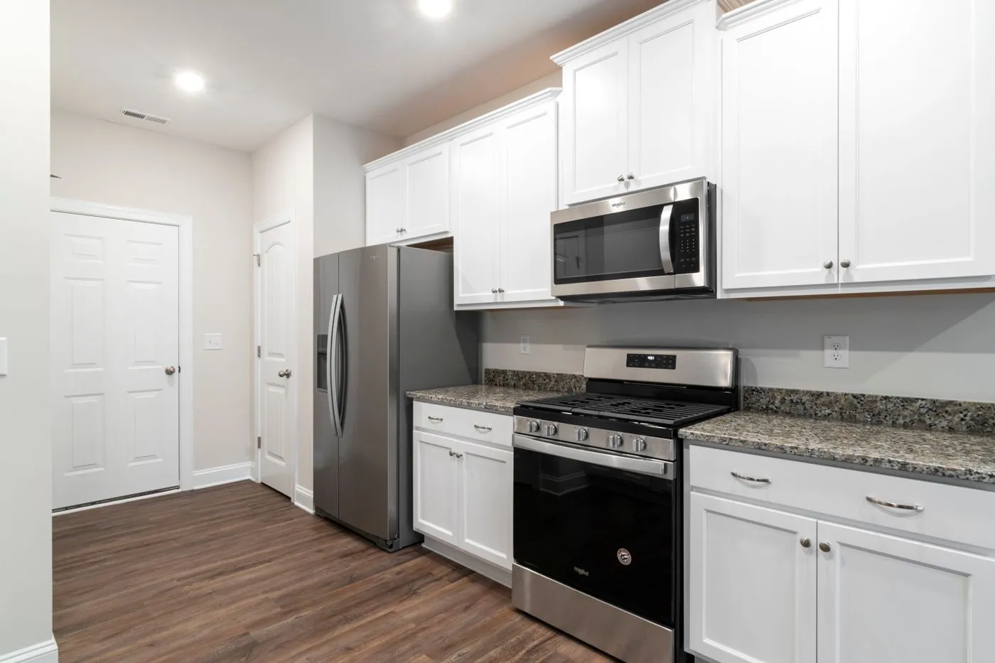 White fitted kitchen with a stainless steel side-by-side fridge beside a doorway and a narrow galley walkway