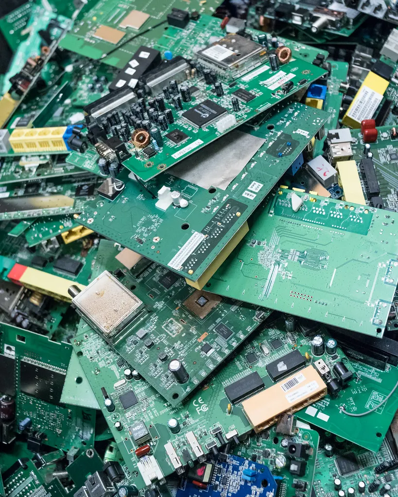 Dense stack of green printed circuit boards from dismantled electronics, showing capacitors, chips, connectors and barcode labels