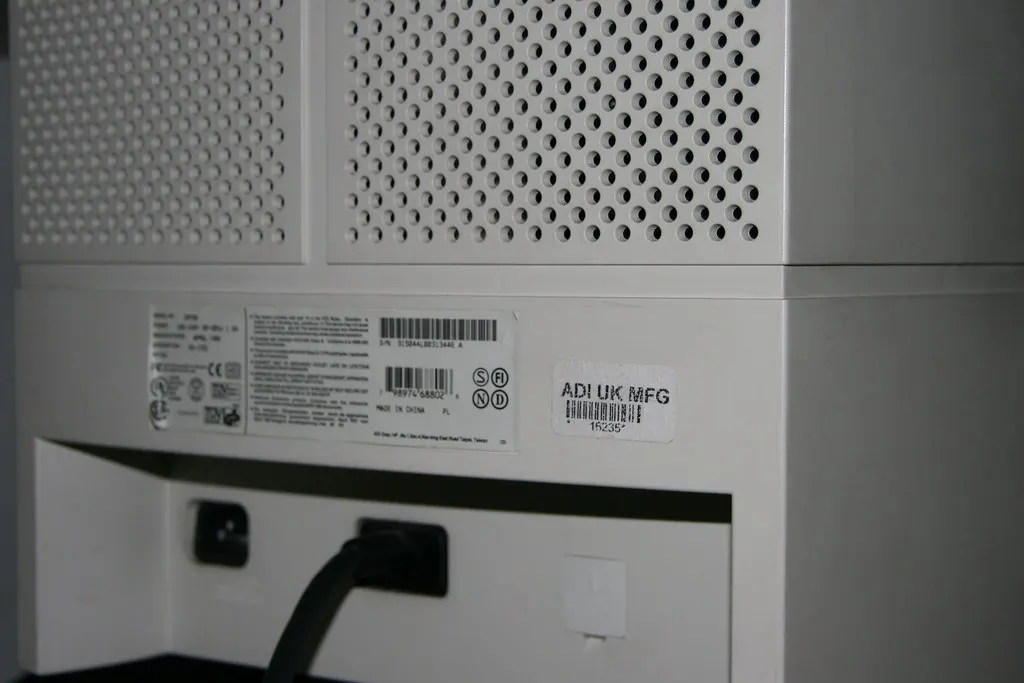 Rear panel of a white desktop computer tower showing a ventilation grille, a power cable socket and printed barcode and serial number labels