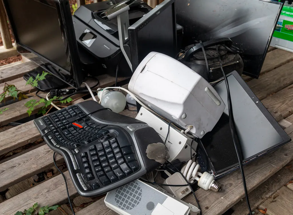 Pile of discarded electronics on a wooden deck including two flat computer monitors, an inkjet printer, a laptop, an ergonomic keyboard and a compact fluorescent bulb