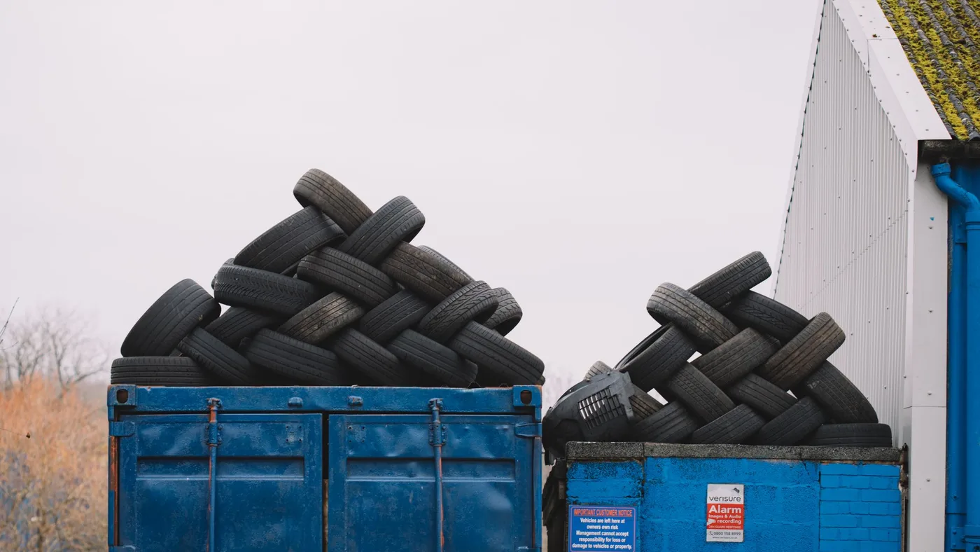 Two blue metal skips heaped with discarded car tyres standing outdoors beside a white wall