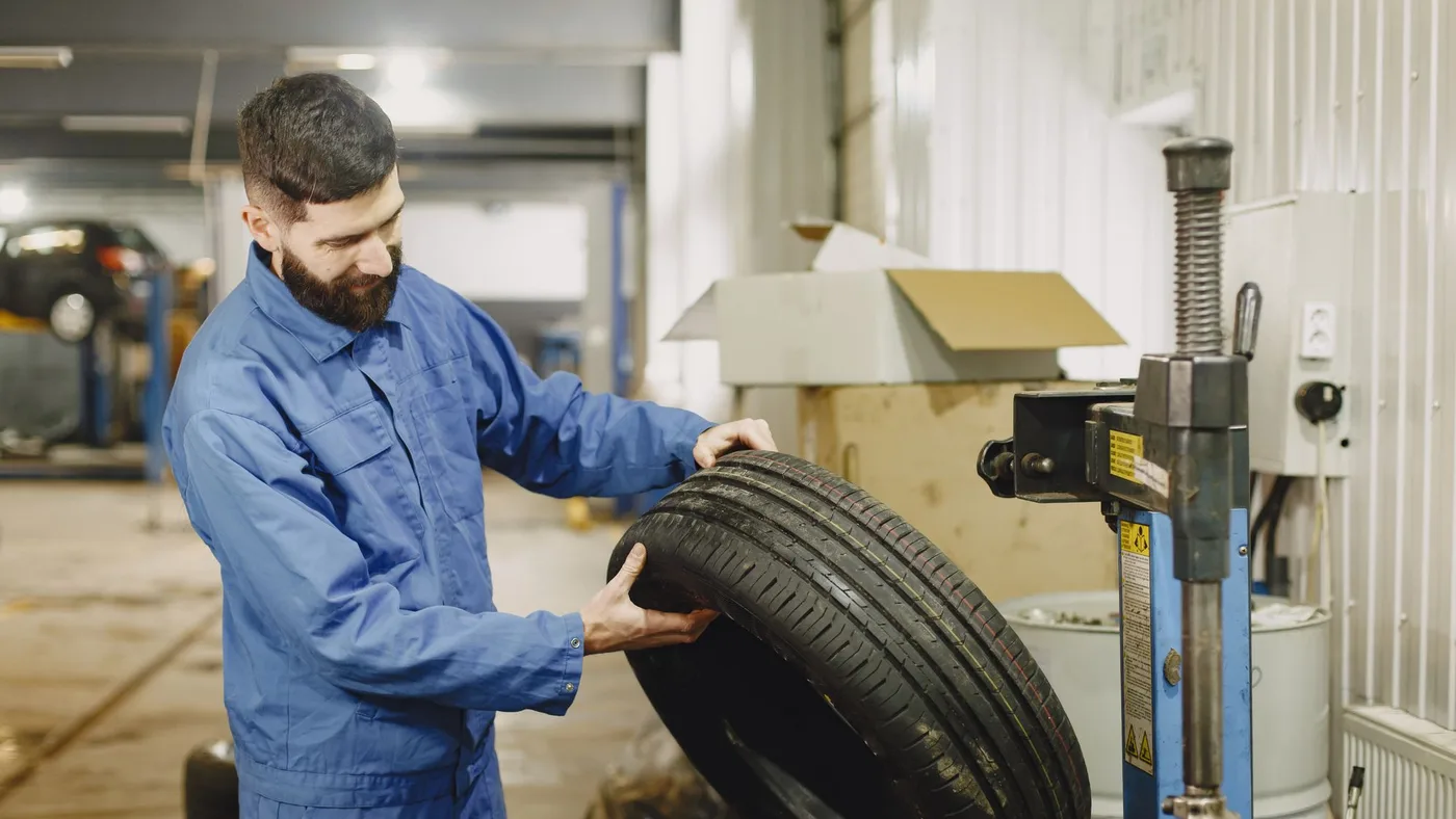 Mechanic in blue coveralls holding a used car tyre upright beside a tyre changing machine in a workshop