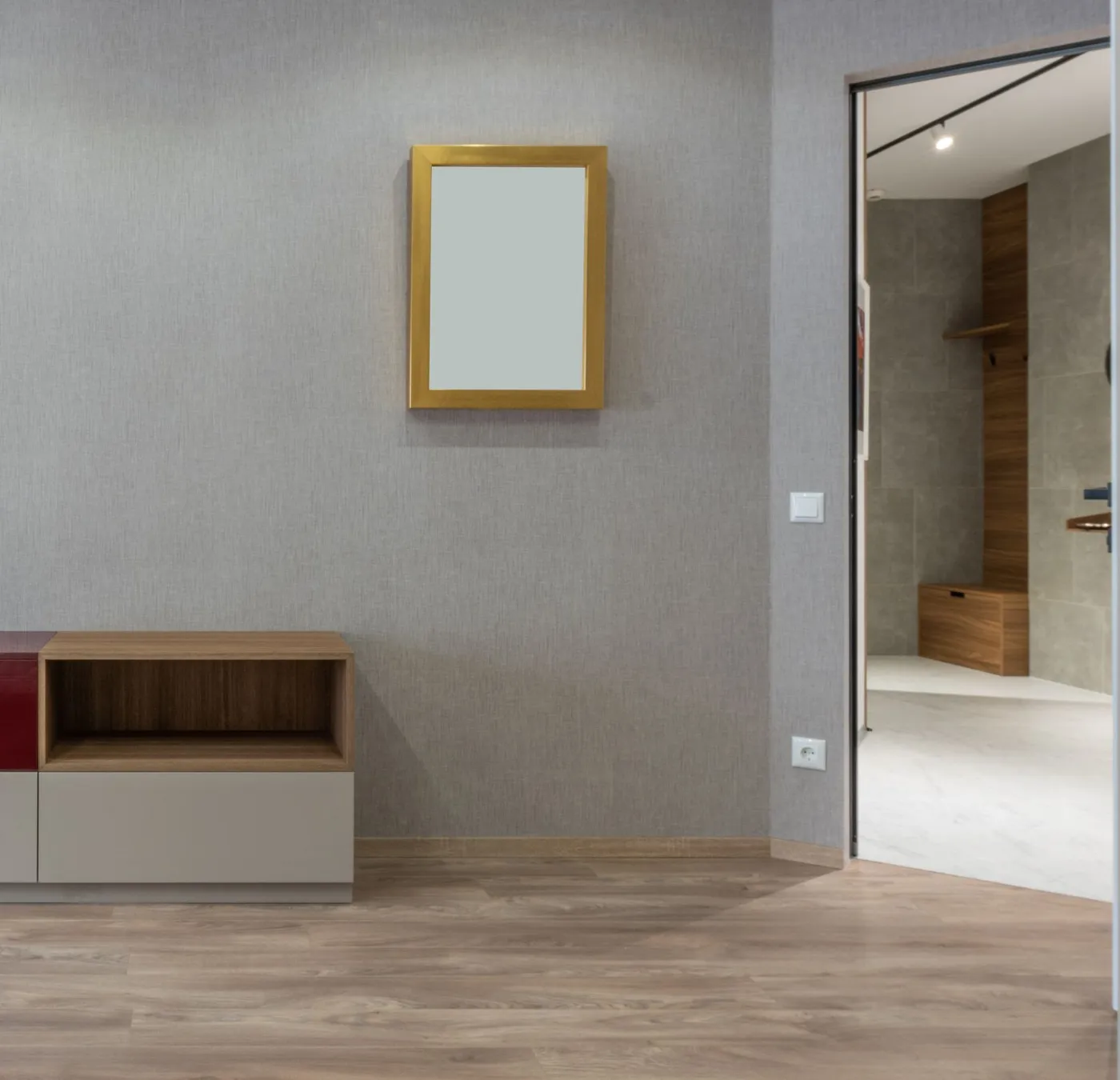 Emptied apartment hallway with a low fitted sideboard, gold-framed mirror and laminate flooring beside an open doorway