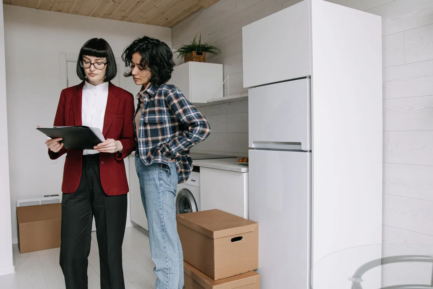 Letting agent in a red blazer holding a clipboard beside a tenant in a kitchen with a fridge, washing machine and packed cartons