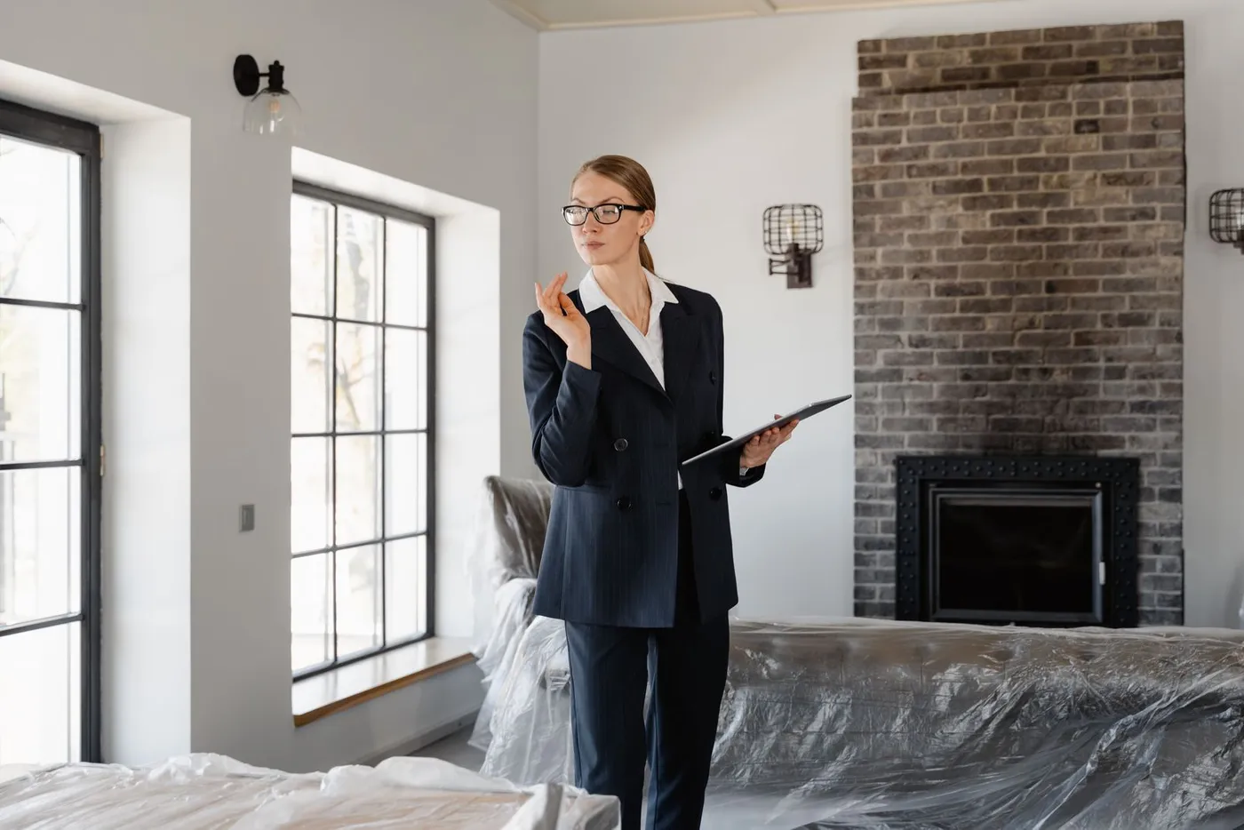 Woman in a dark suit holding a tablet while walking through a bare room with furniture covered in protective sheeting