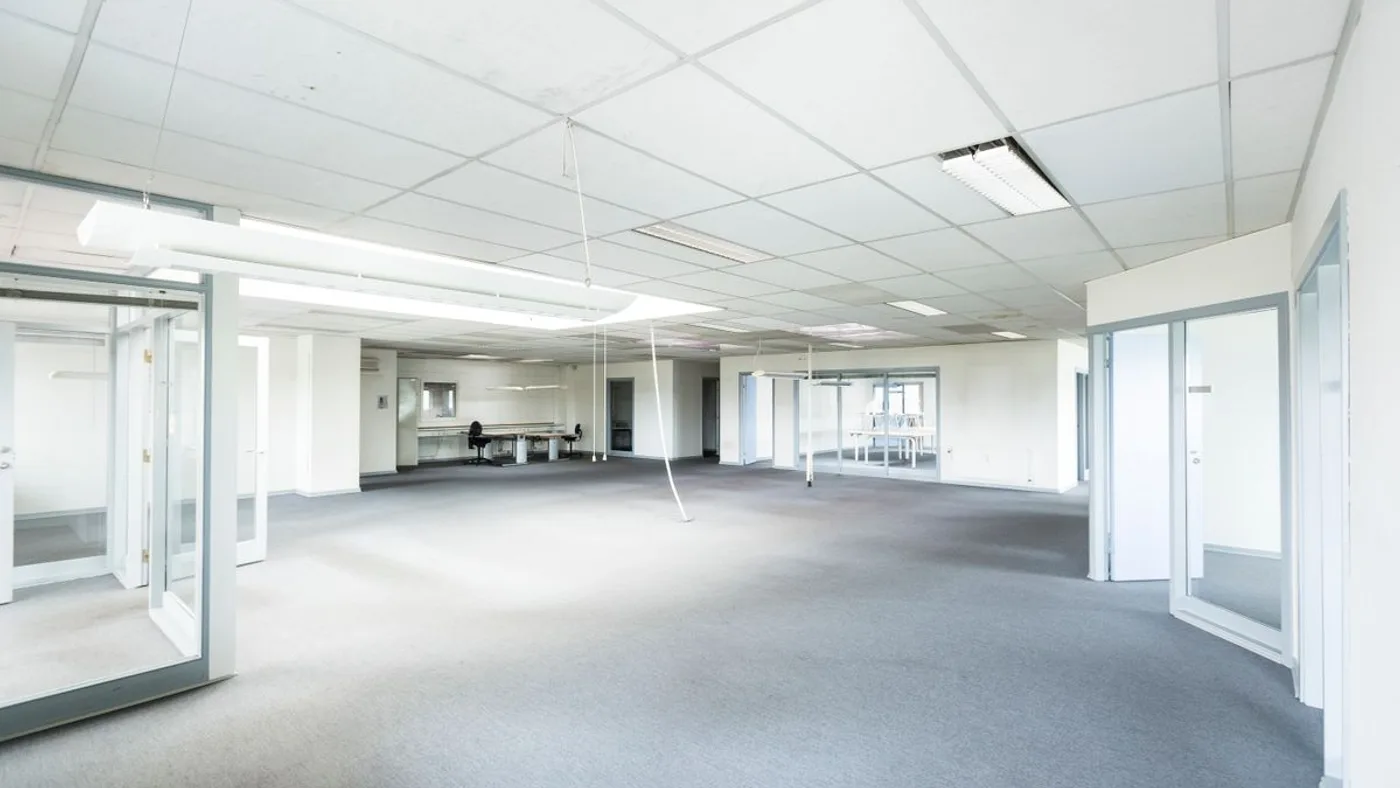 Emptied office floor with grey carpet, suspended ceiling tiles, strip lighting and glass partition doors along both sides