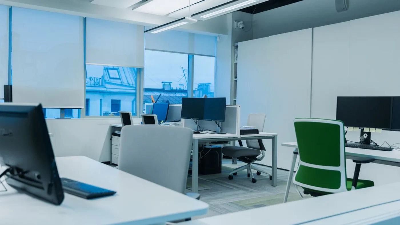Open-plan office with white desks, dual monitors, grey and green task chairs and mobile pedestal units behind partition screens