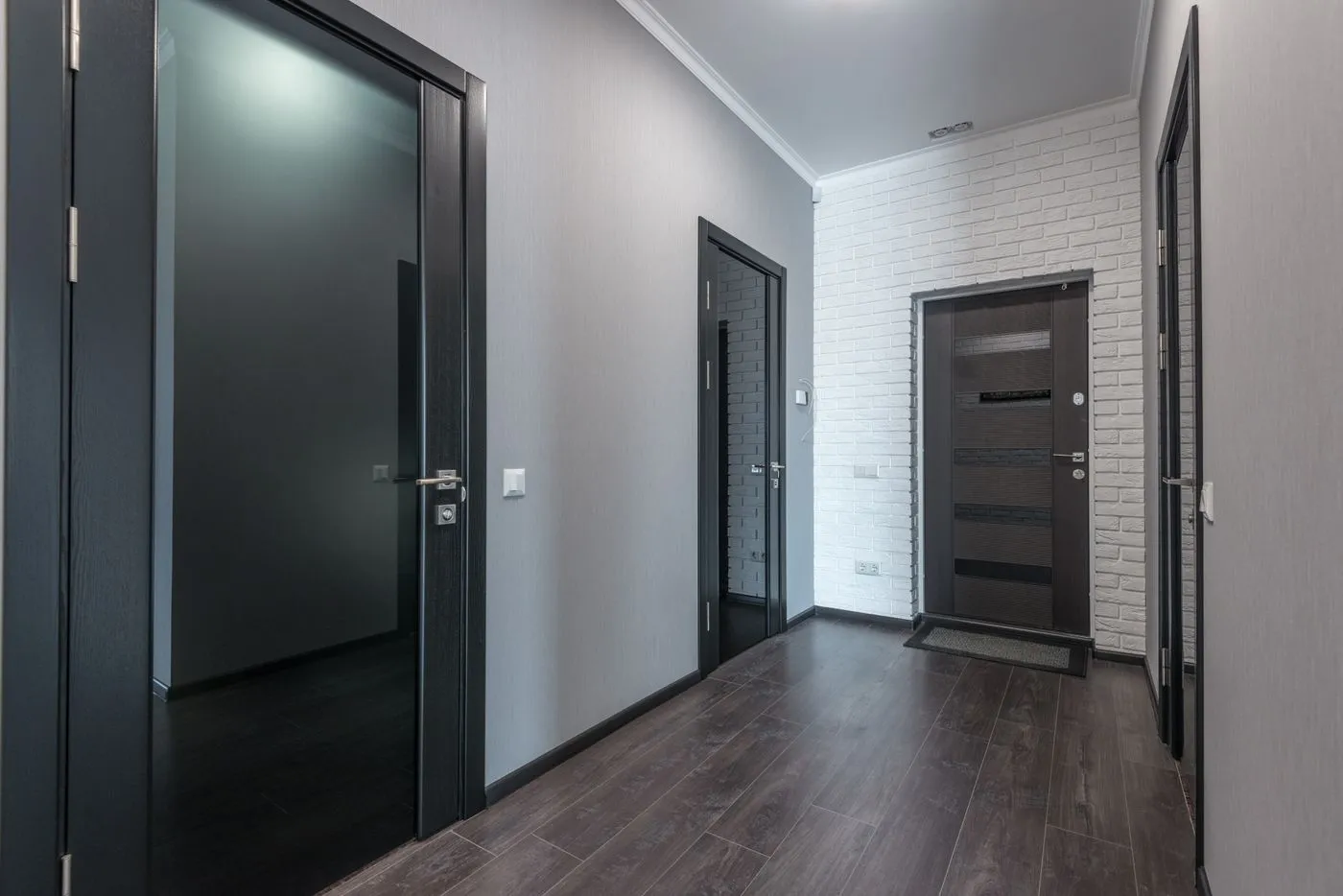 Empty modern apartment hallway with dark wood doors, grey walls and a white brick accent wall leading to a front door