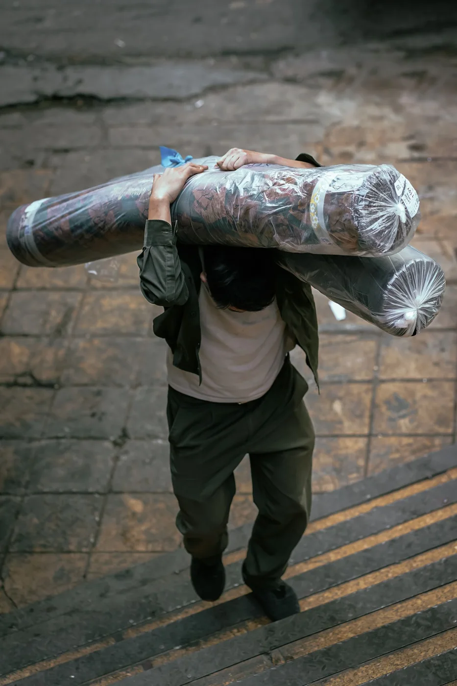 Person in a dark green jacket carrying two long plastic-wrapped rolls balanced across one shoulder while walking up wet stone steps