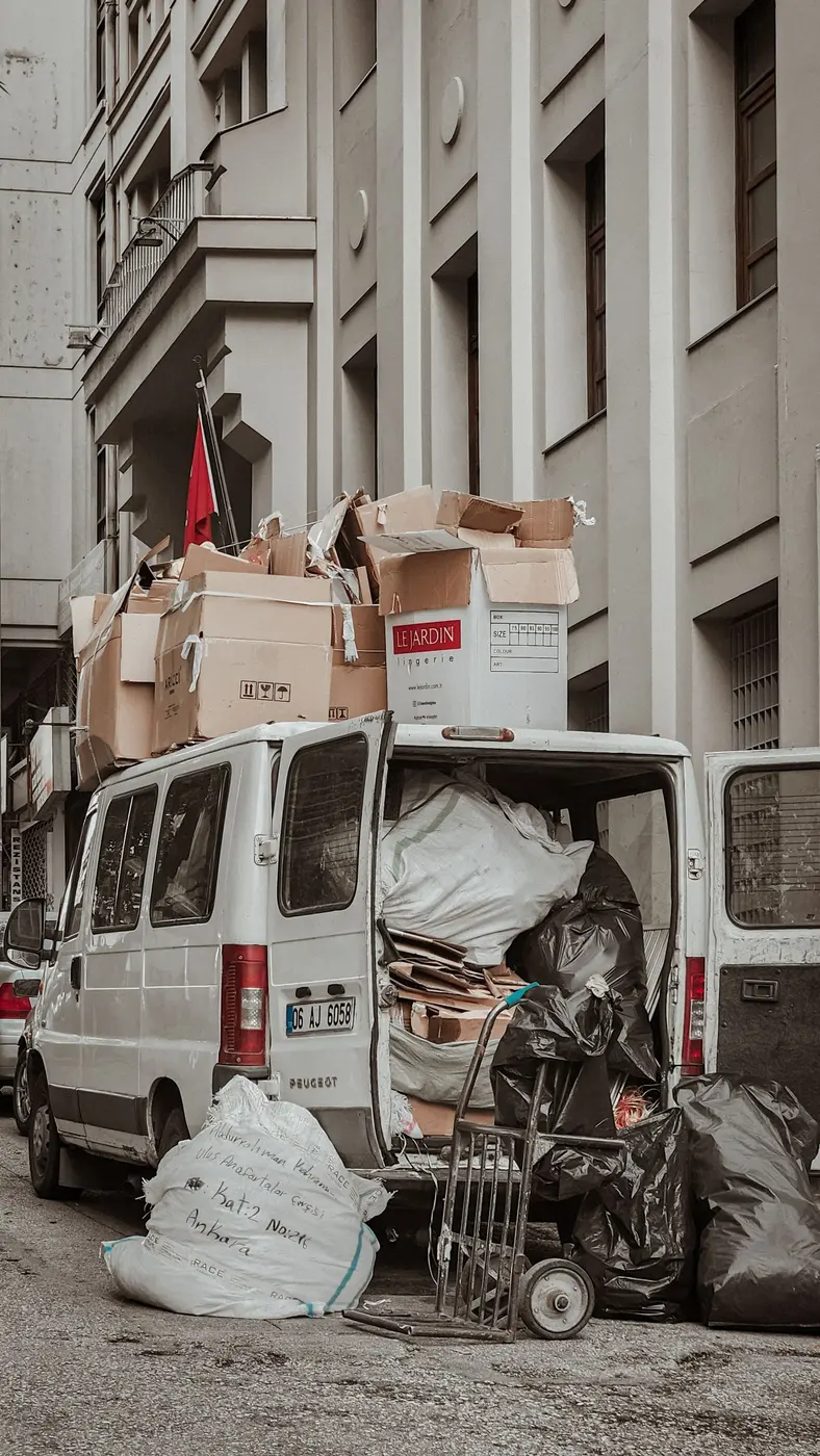 Small white van packed with sacks, flattened cardboard and black bags, more boxes stacked on its roof and a sack trolley standing on the pavement beside it