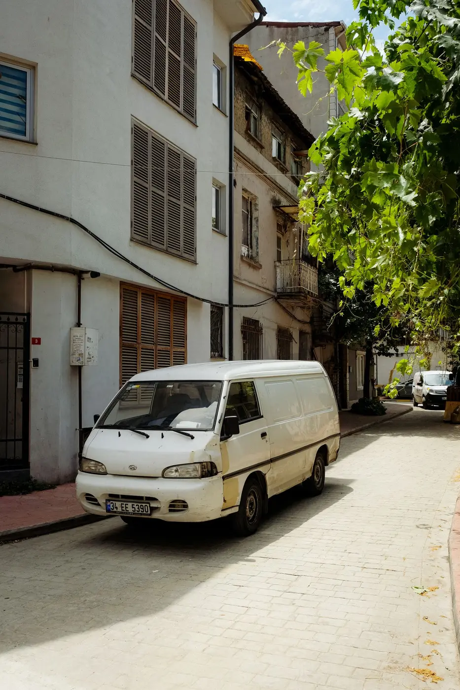 White panel van parked tight against the kerb on a narrow paved residential street, with a second van waiting further down between the buildings