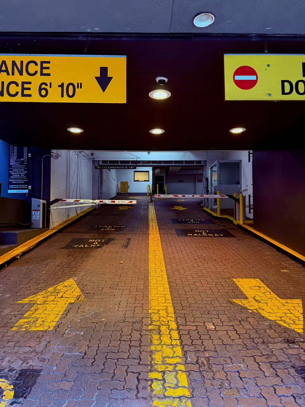 Covered car park entrance with a yellow overhead clearance height sign, raised barrier arms and yellow arrows painted on the brick ramp