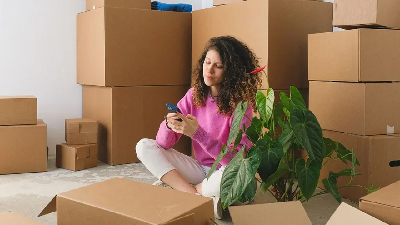 Woman in a pink jumper sitting cross-legged on the floor checking her phone among stacked cardboard boxes and a potted plant