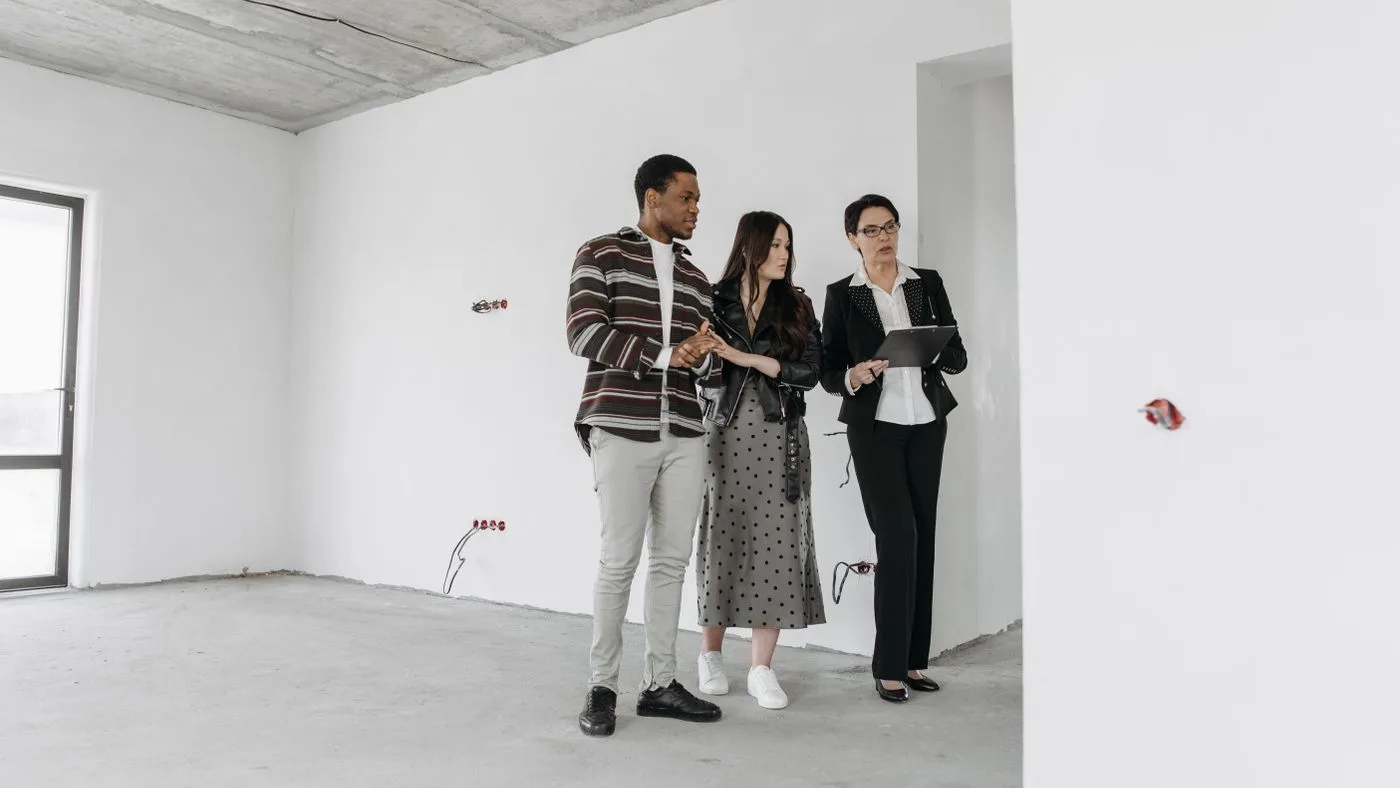 Three people standing with a tablet in a bare, unfurnished apartment with white walls and an untreated concrete floor