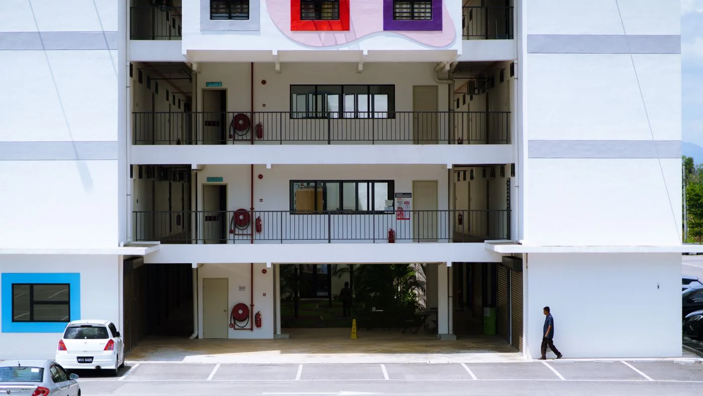 Facade of a low-rise residential block showing three floors of open walkways with railings, above a row of marked parking bays