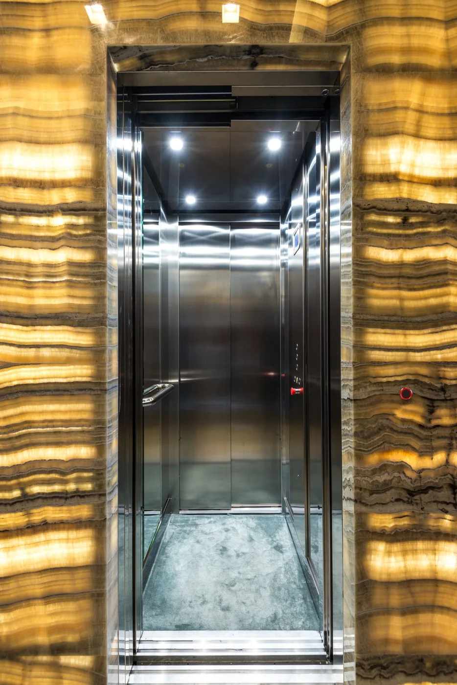 Stainless steel lift car with open doors set into a backlit onyx stone wall in a residential building lobby