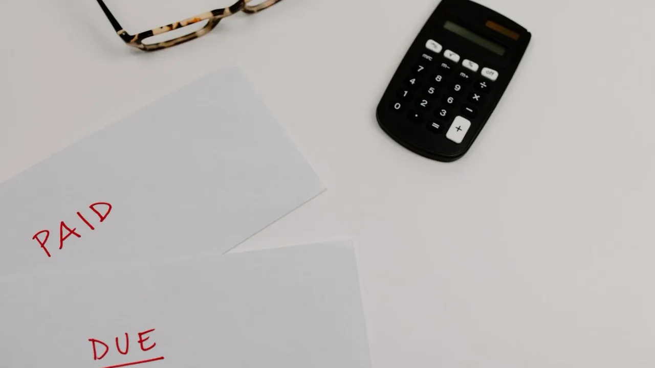 Two sheets of white paper marked PAID and DUE in red ink beside a black calculator and a pair of glasses on a pale desk