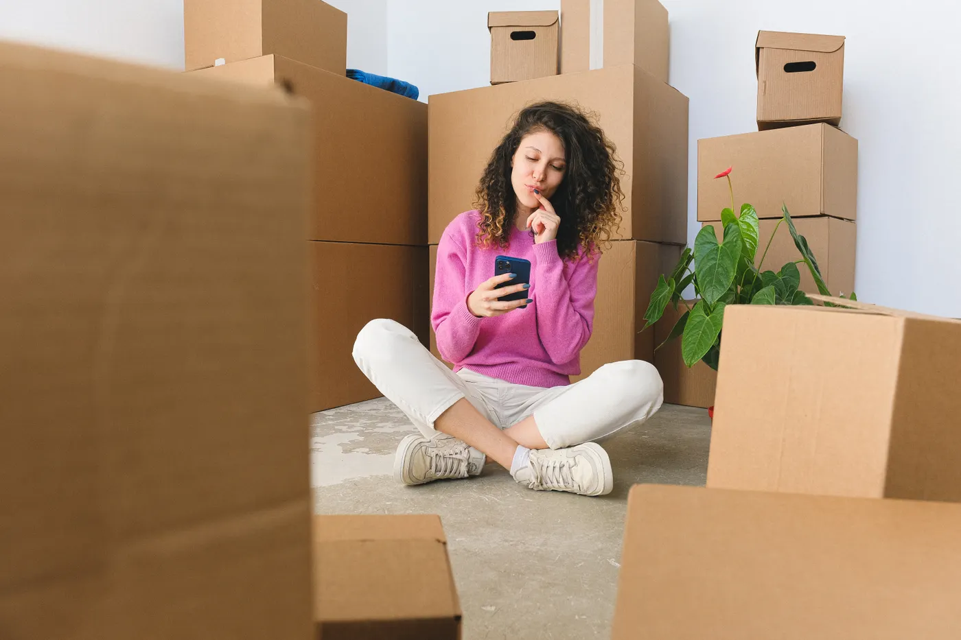 A woman sitting among moving boxes, smiling while messaging on a smartphone to arrange a junk removal collection in Dubai