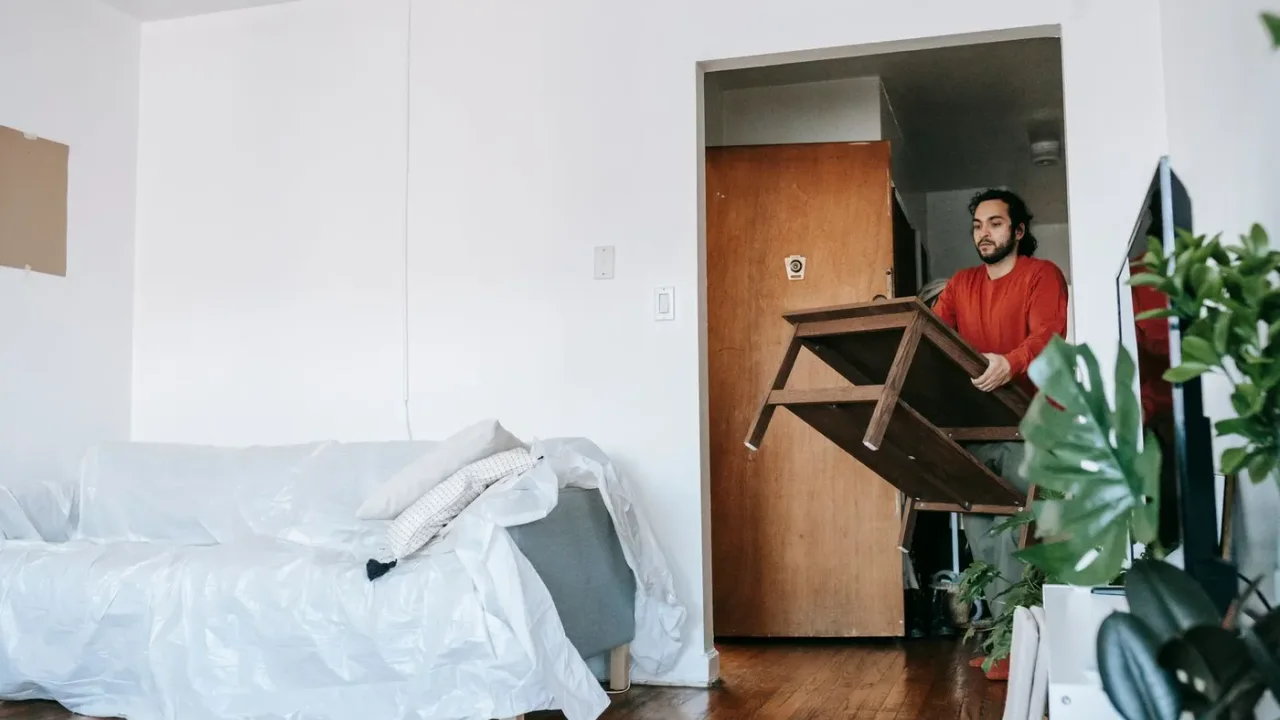 Man in a red sweater tilting a dark wooden table through an apartment doorway, with a sofa under a plastic dust sheet in the foreground