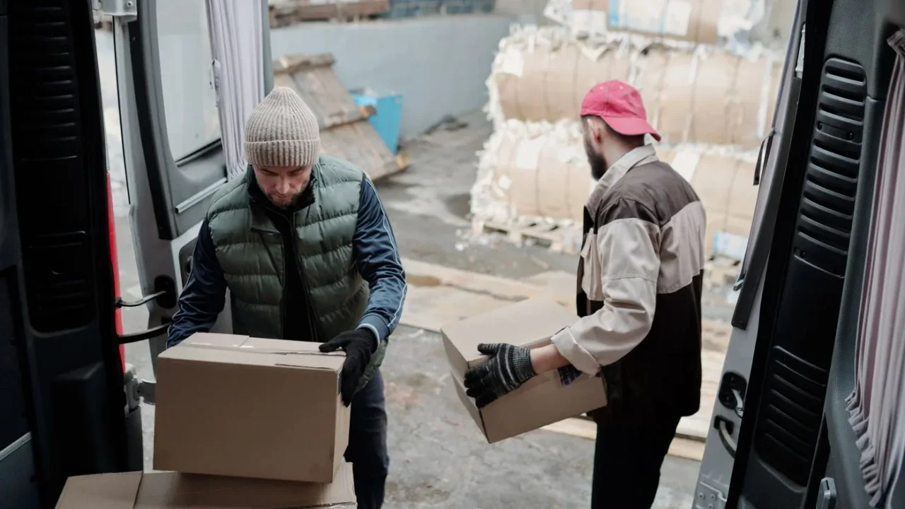 Two workers in beanies and work gloves loading cardboard boxes into the open rear doors of a van, with wrapped bales stacked behind them
