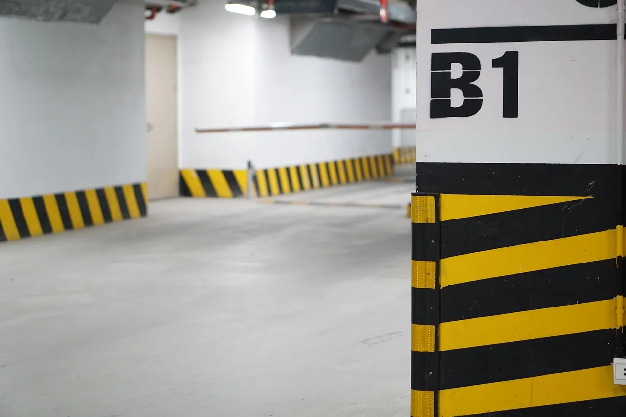 Empty basement car park level marked B1 on a white column with yellow and black hazard stripes and a low concrete ceiling with ducting