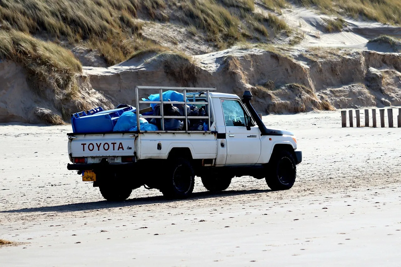 White Toyota pickup truck loaded with blue crates and bagged waste behind a raised rear frame, parked on pale sand beside dunes