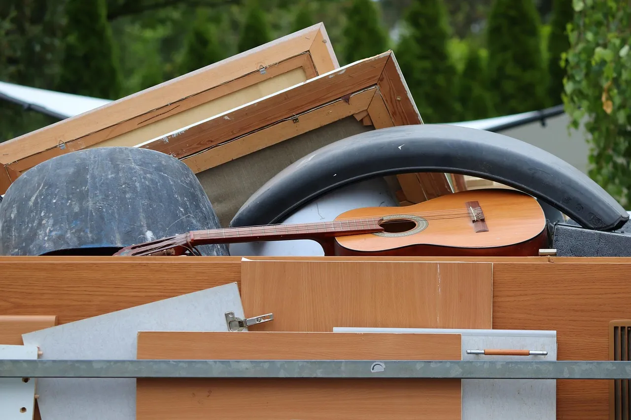 Loaded open truck bed piled with timber window frames, chipboard panels, a plastic tub, a car mudguard and an acoustic guitar stacked above the side rails