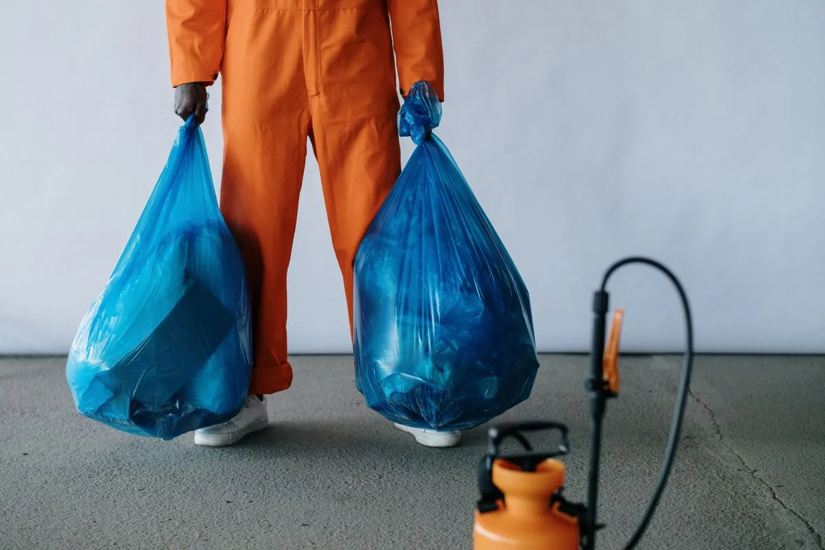 Worker in orange overalls carrying two full blue waste bags across a concrete floor