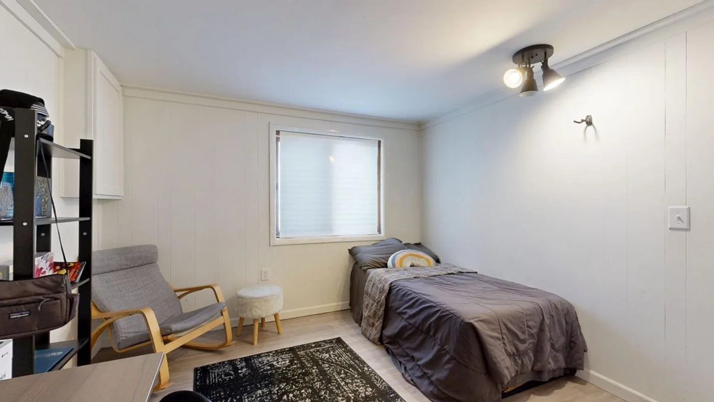 Small plain room with a single bed against a white panelled wall, a wooden rocking chair, a low shelf unit and a dark patterned rug