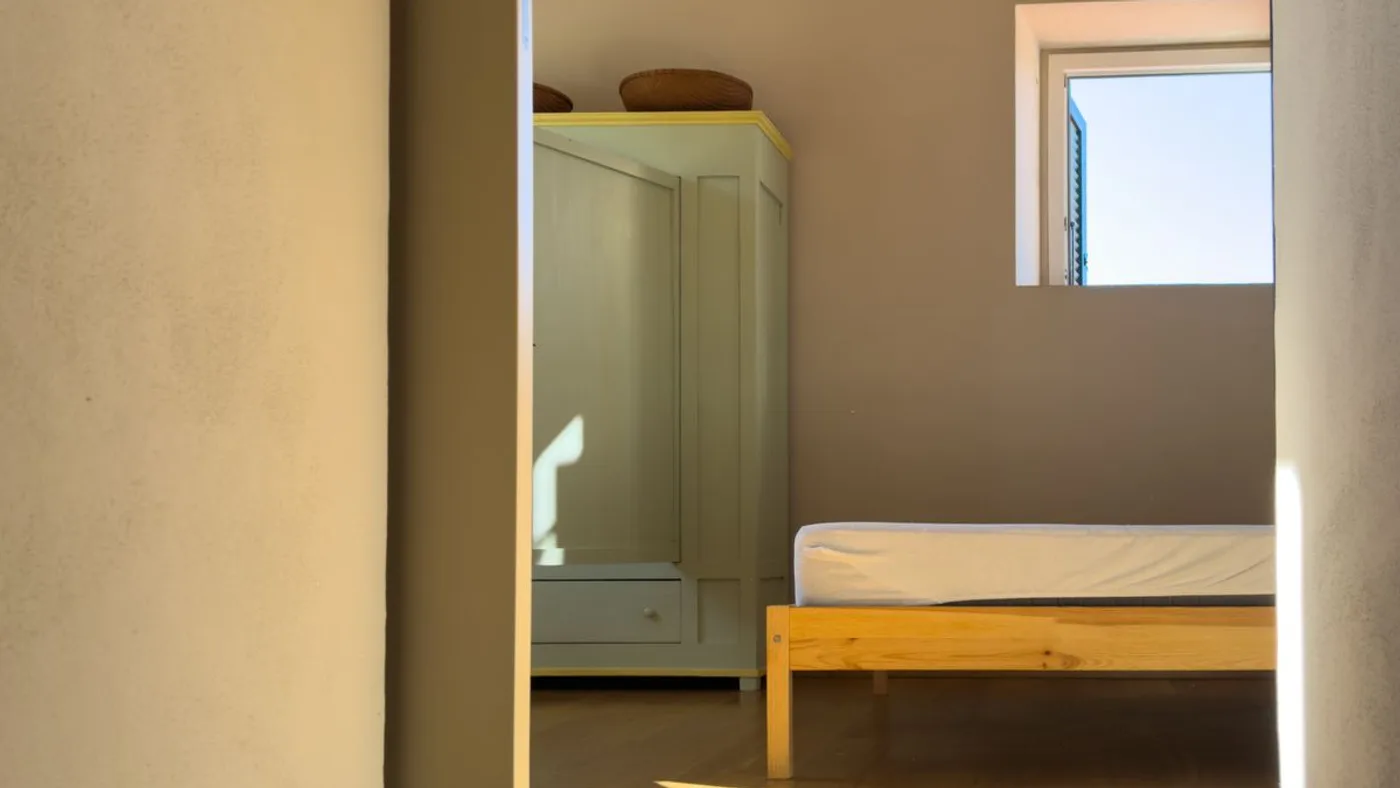 View through a narrow open doorway into a small sunlit room containing a plain wooden bed frame and a pale green freestanding wardrobe