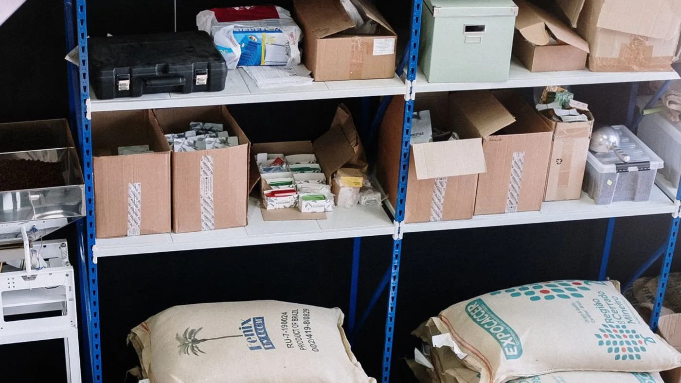 Metal shelving racks in a small utility room packed with cardboard boxes, plastic cases and stacked sacks with more items on the floor beneath