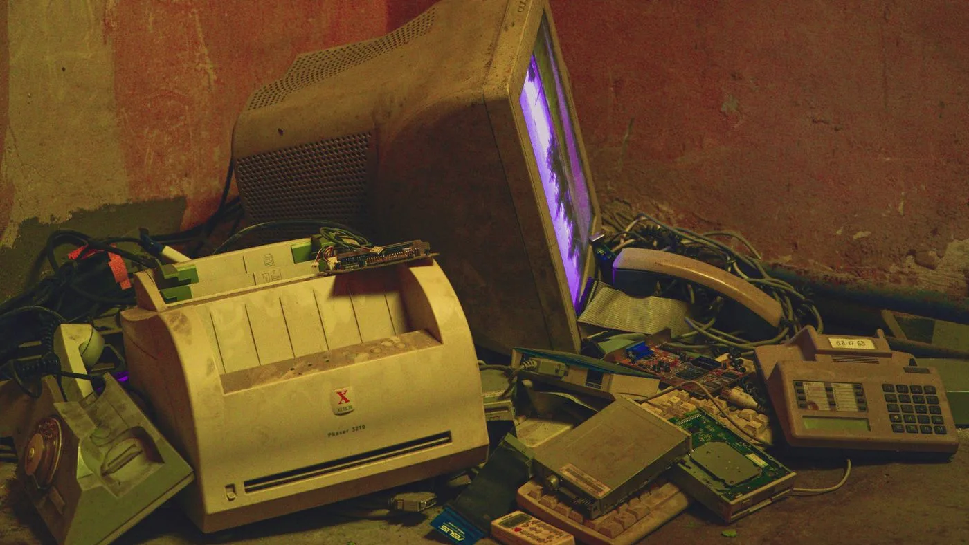 Pile of discarded electronics against a wall including a beige CRT monitor, a desktop printer, a telephone, circuit boards and tangled cables