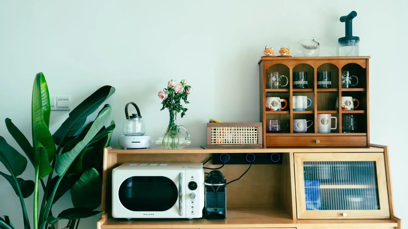 Cream countertop microwave on a wooden sideboard beside a coffee machine, a glass kettle and a wall cabinet of mugs
