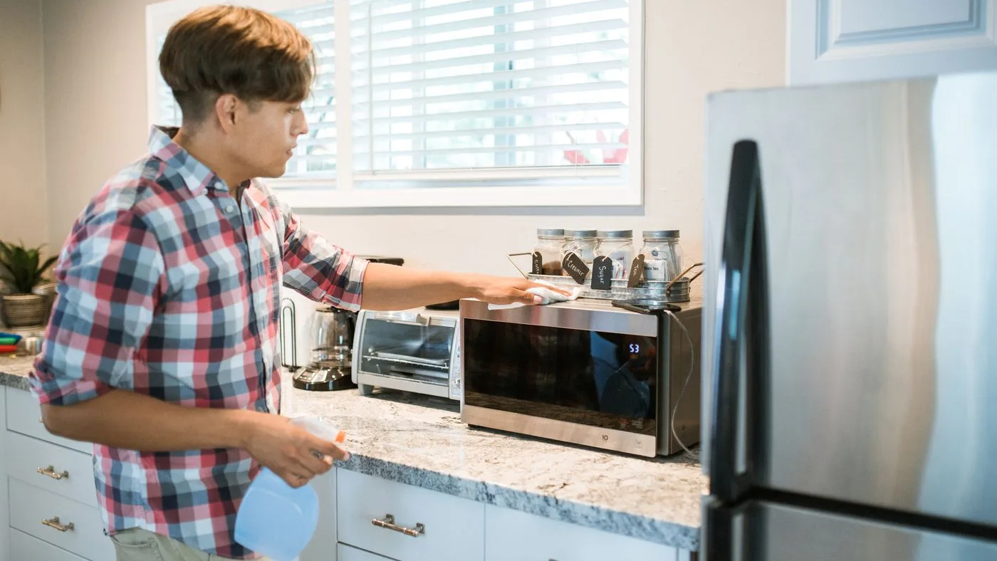 Person in a checked shirt wiping the front of a stainless steel microwave on a granite worktop, holding a spray bottle