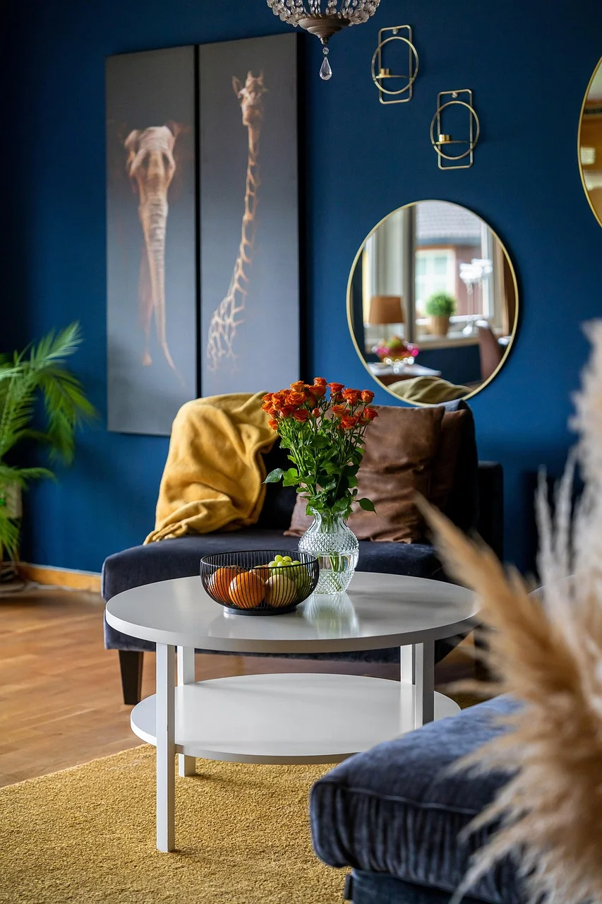 Round gold-framed wall mirror hanging on a deep blue wall above a velvet bench, with a white round side table and a vase of red roses in front