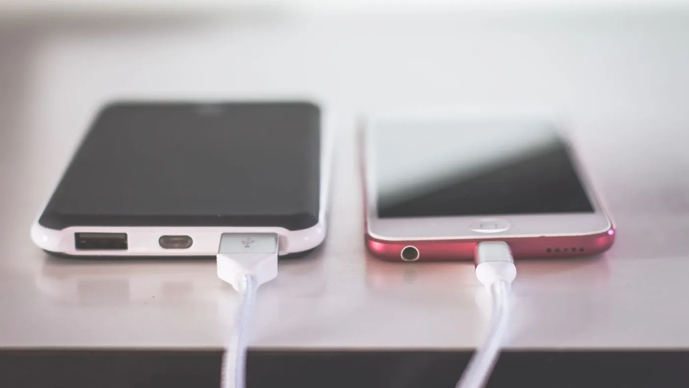 Two smartphones lying on a pale surface with white charging cables plugged into their ports