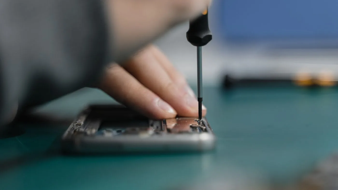 Hand using a precision screwdriver on an opened phone chassis resting on a green work mat