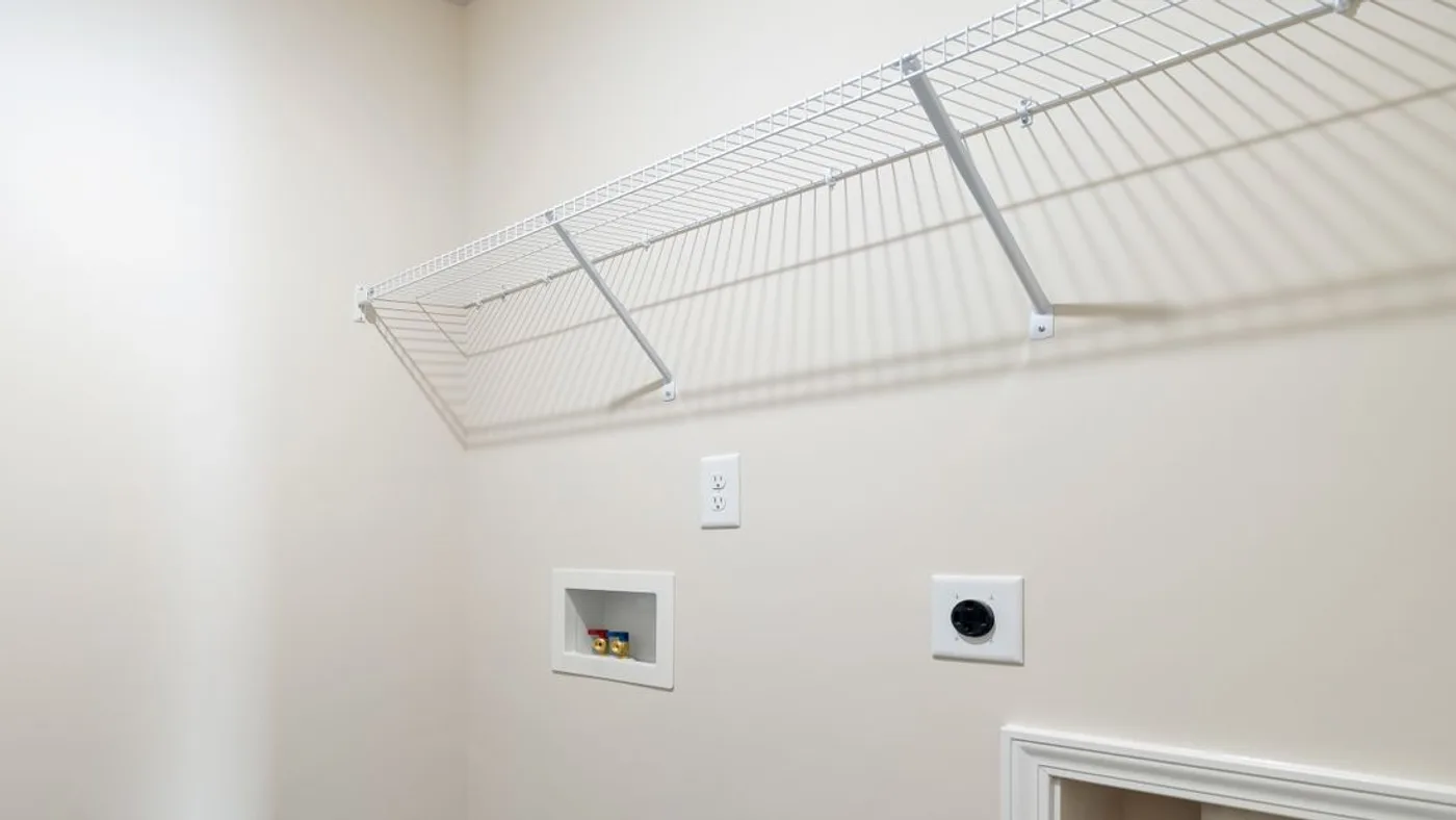 Empty laundry alcove with a recessed water valve box, a power socket, a vent duct opening and a white wire shelf above