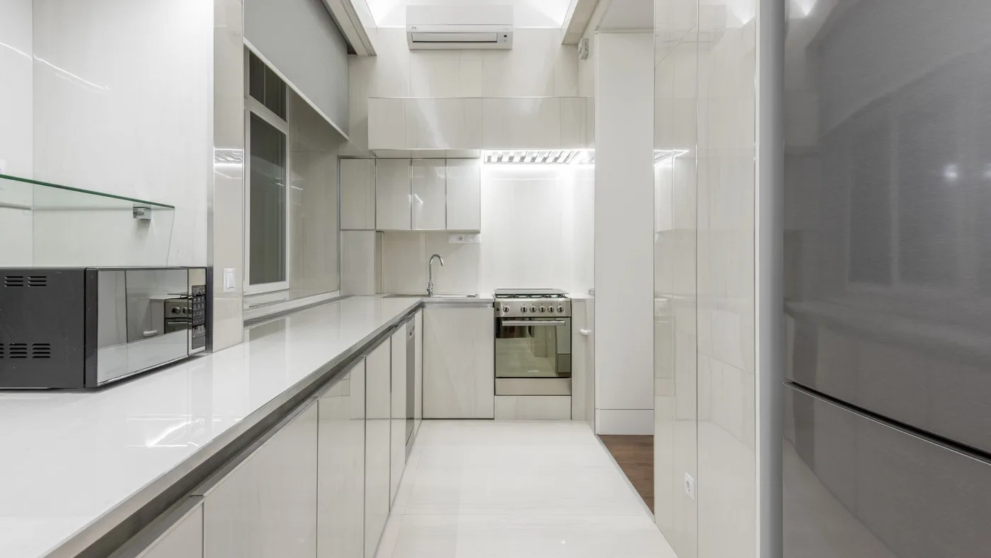 Narrow white gloss galley kitchen with a wall-mounted split air conditioning unit, freestanding gas cooker and tall stainless fridge