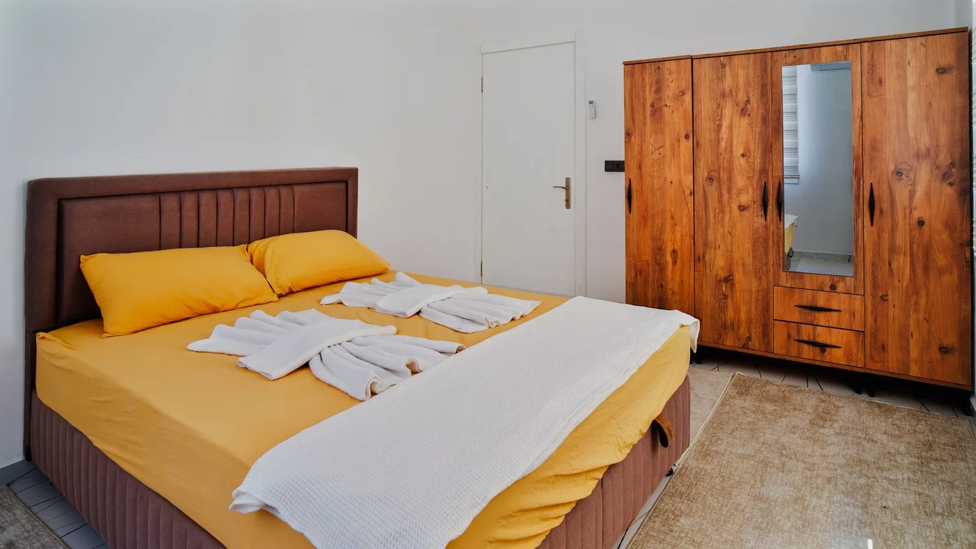 Furnished bedroom with a made double bed, folded towels and a tall free-standing wooden wardrobe with a mirrored door