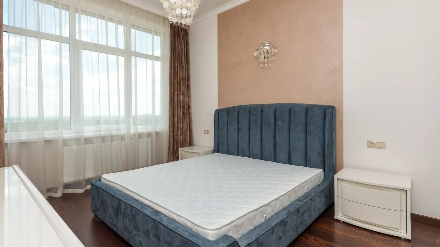 Stripped bare mattress on a blue upholstered bed base beside two white bedside cabinets in a cleared bedroom