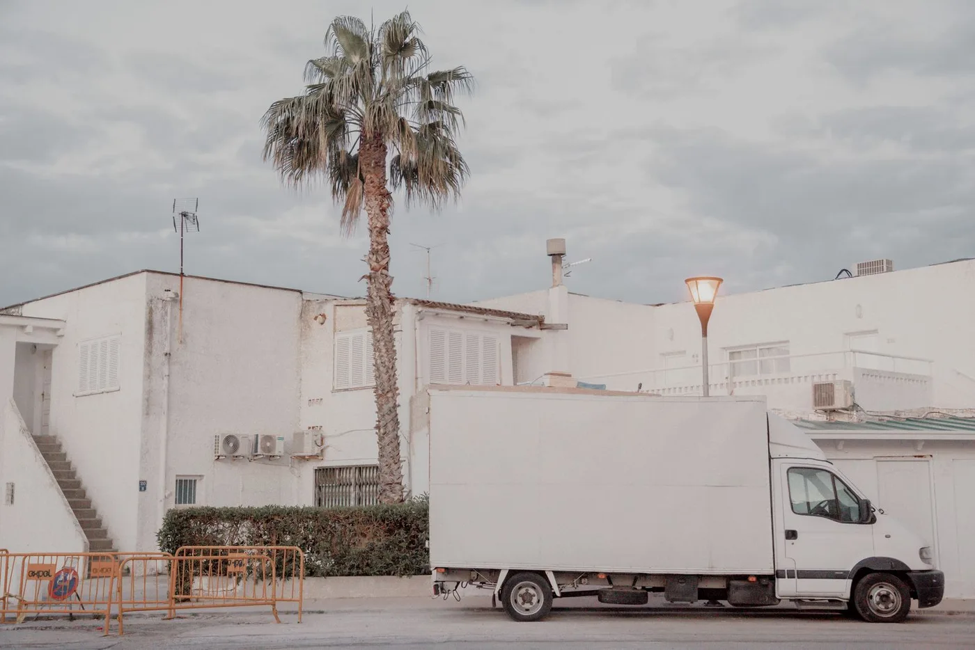 White box removal truck parked on the street beside a plain white building with a palm tree overhead