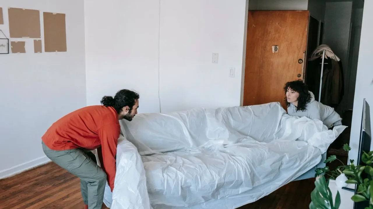 Two people bending to lift a sofa wrapped in a white dust sheet across the wooden floor of an apartment living room