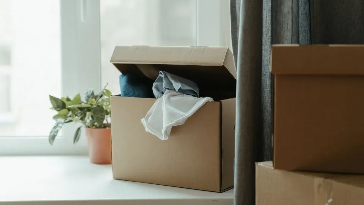 Open cardboard box of folded clothes on a white windowsill beside a potted plant, with more boxes stacked by a grey curtain