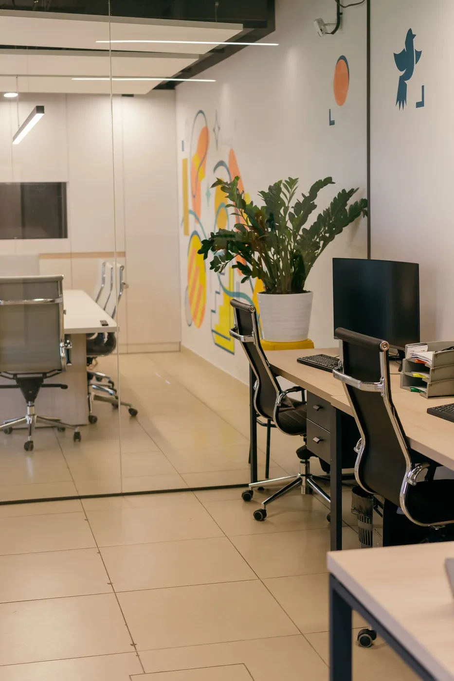 Office with light wood desks, black leather chairs, a monitor and a glass-partitioned meeting room behind