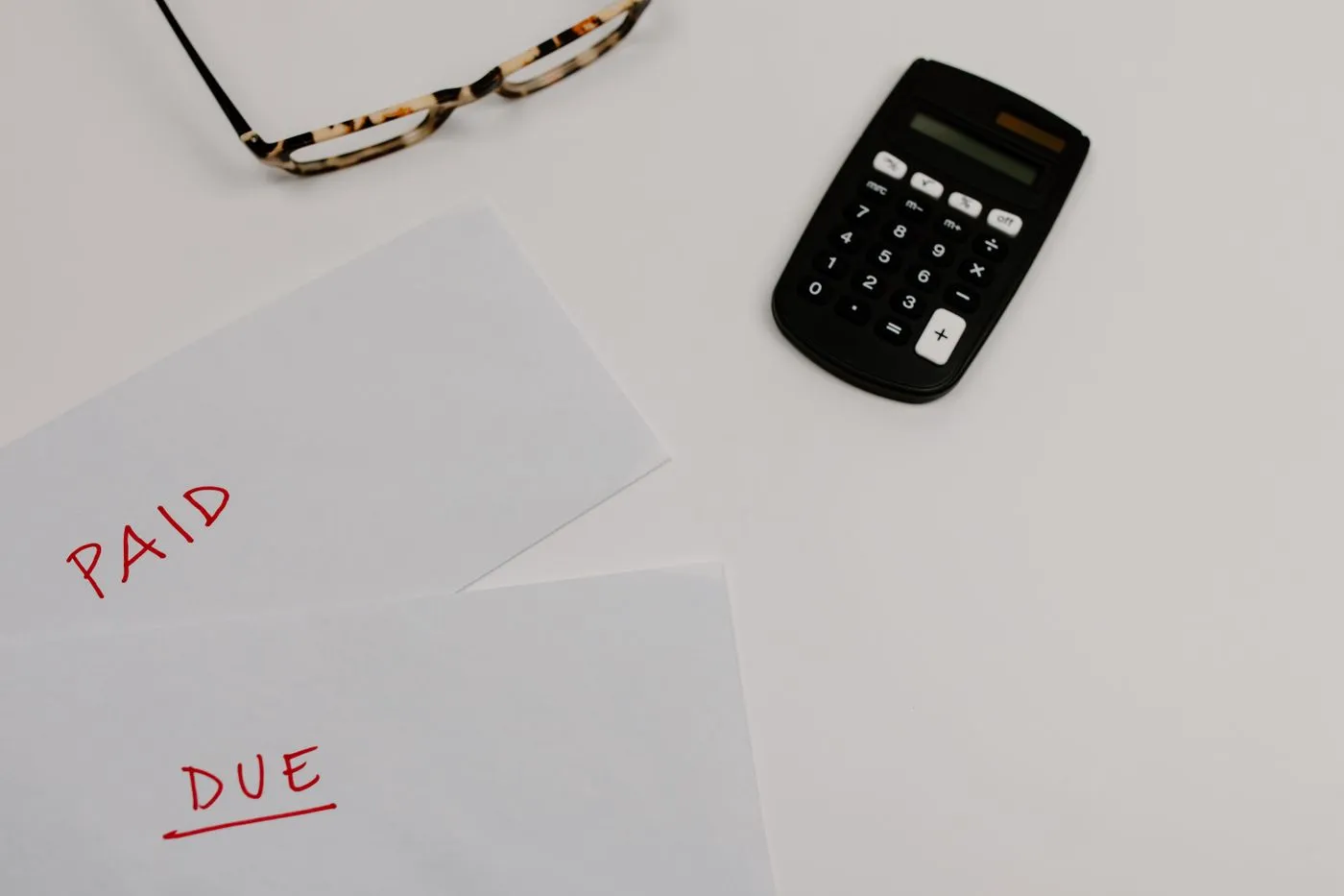 Two white envelopes marked PAID and DUE in red beside a black calculator and tortoiseshell reading glasses