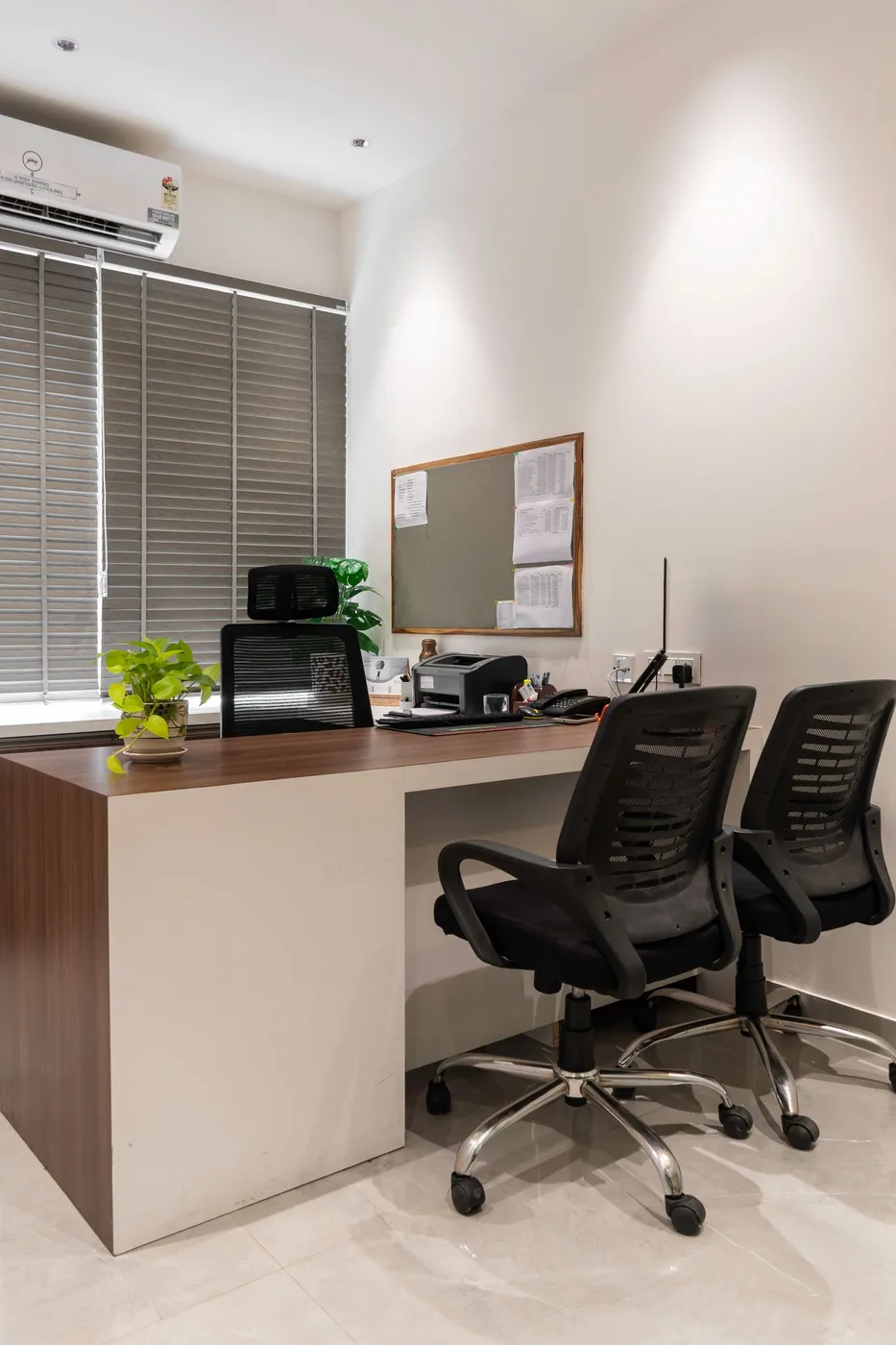 Small private office with a fitted wooden desk, two black mesh chairs, a printer, noticeboard and wall-mounted air conditioner
