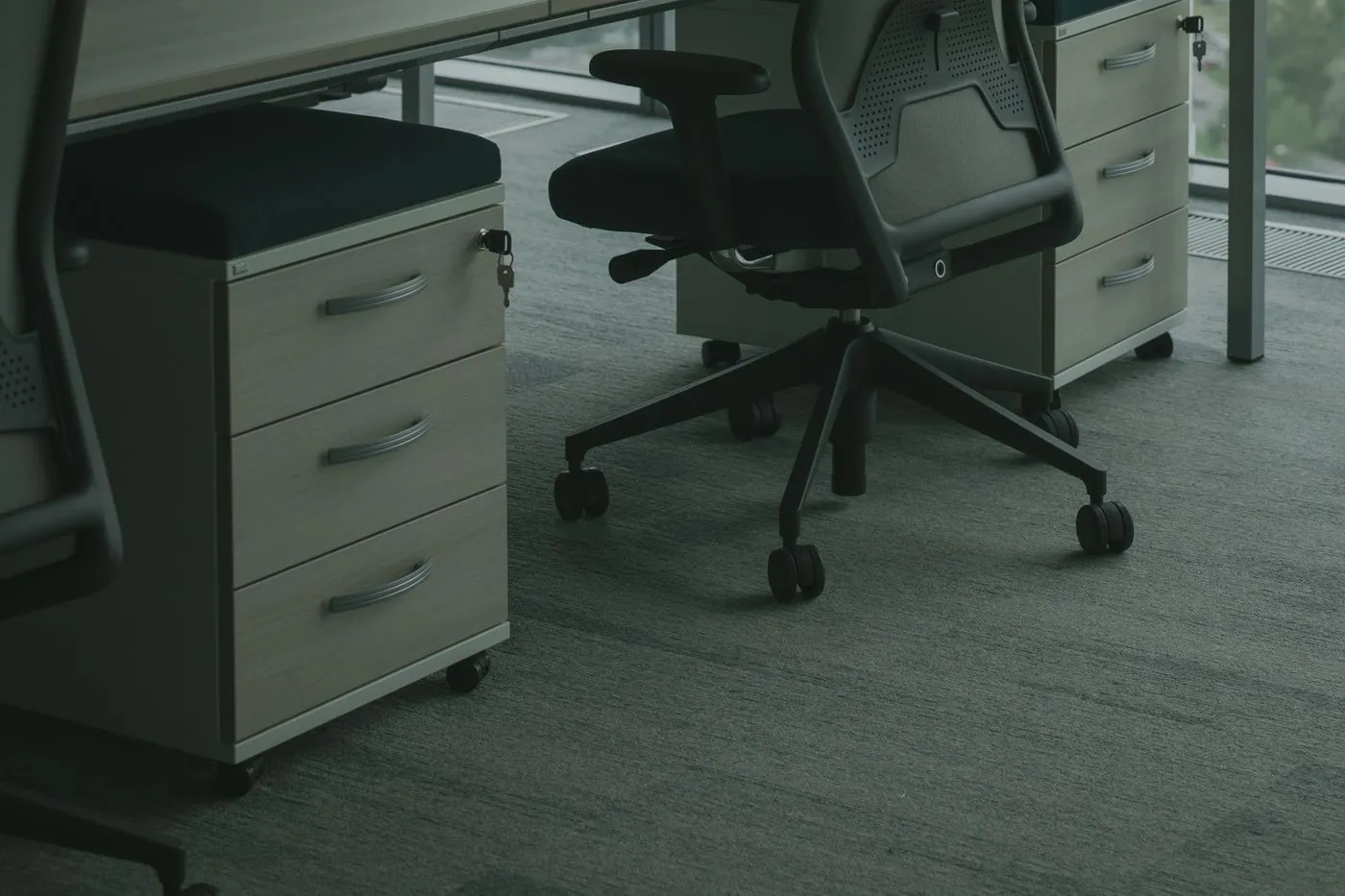 Lockable wooden office pedestals with keys in the locks beside a black task chair on grey carpet tiles
