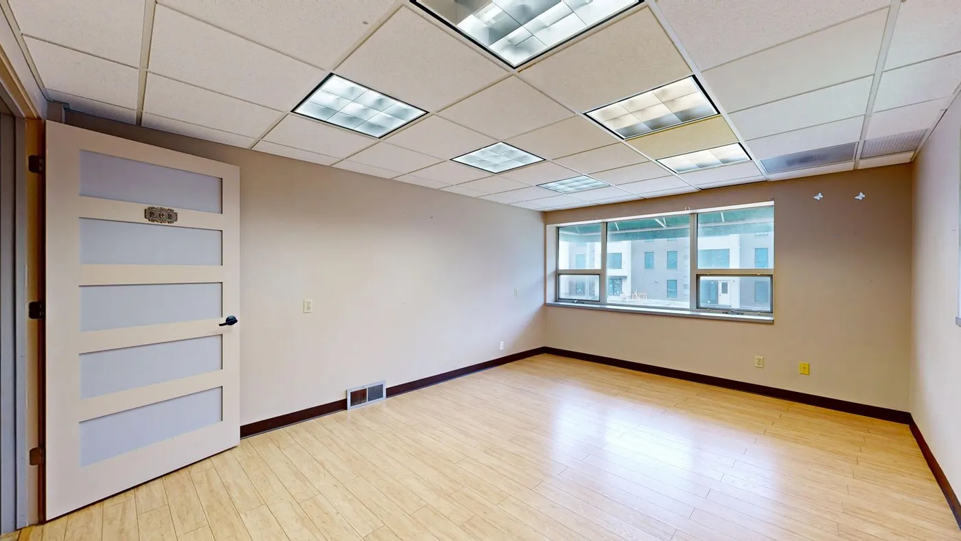 Small empty office room with pale wood flooring, a drop ceiling and a window overlooking a construction site